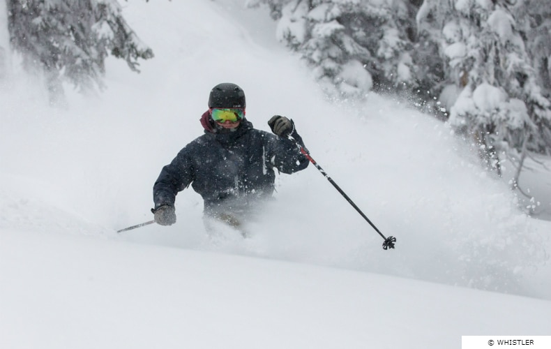 A skier in black with mirrored visor and ski poles navigates some fresh powder snow