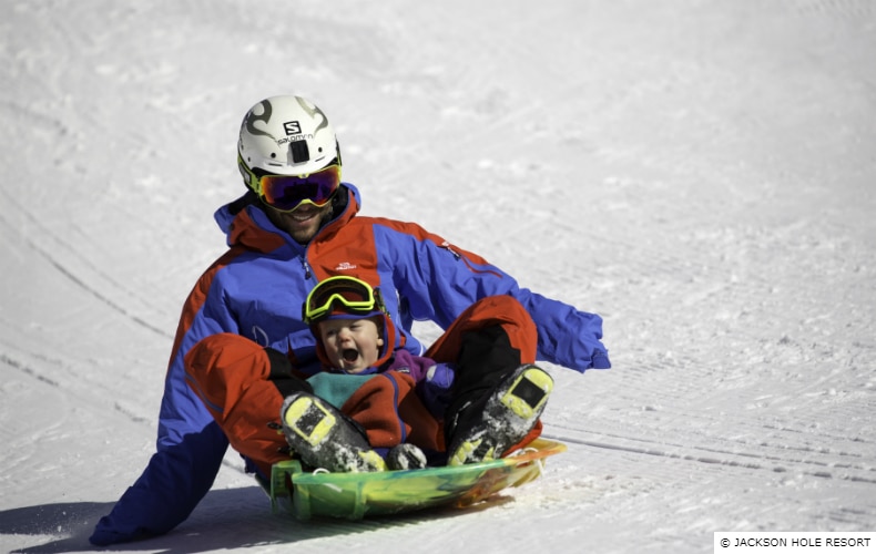 A father and young child laughing as they slide down a slope on a plastic sled 