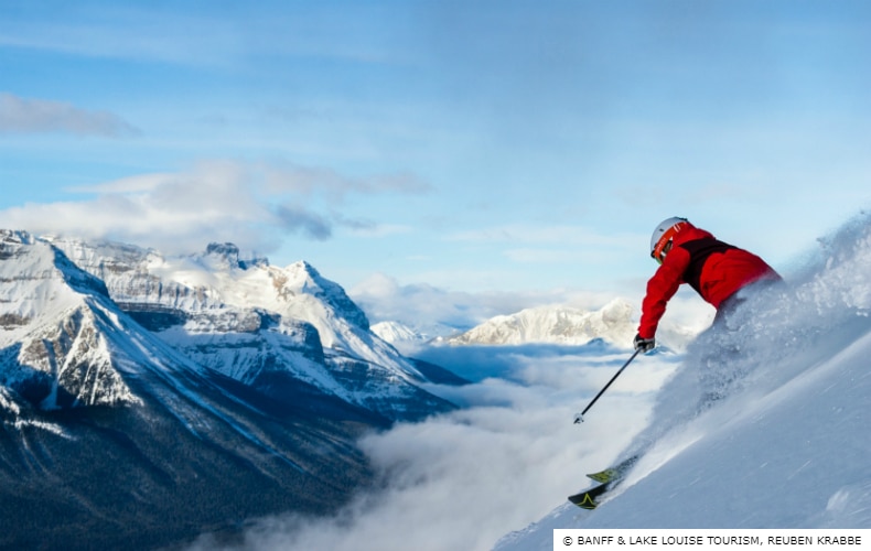 A skier in red at Lake Louise Ski Resort descends the mountain with blue skies and Canada's Rocky Mountains in the background